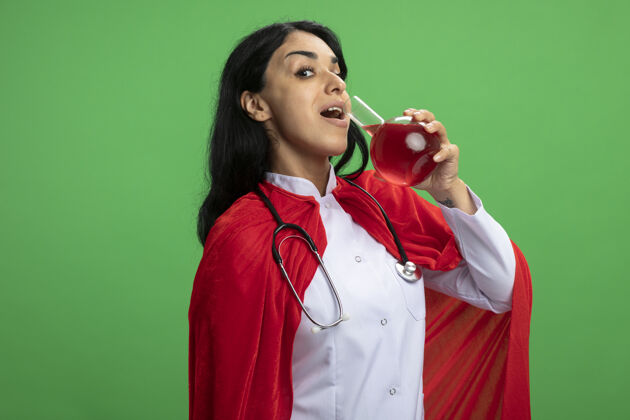 长袍高兴的年轻超级英雄女孩穿着医用长袍听诊器举行和饮料化学玻璃瓶装满红色液体隔离在绿色年轻穿化学