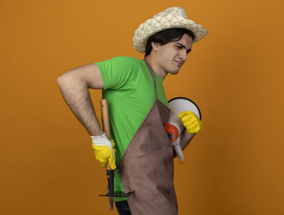 请高兴的年轻男园丁穿着制服戴着园艺帽戴着手套拿着扩音器用耙子刮着腰间的橘子园艺园艺帽子