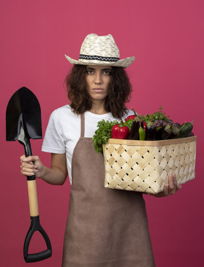 粉红色自信的年轻女园丁 穿着制服 戴着园艺帽 手里拿着蔬菜筐 铁锹插在粉红色的地板上黑桃制服花园
