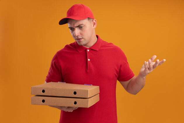 男人困惑的年轻送货员穿着制服 戴着帽子 看着披萨盒摊开 手被隔离在橙色的墙上制服拿着盒子