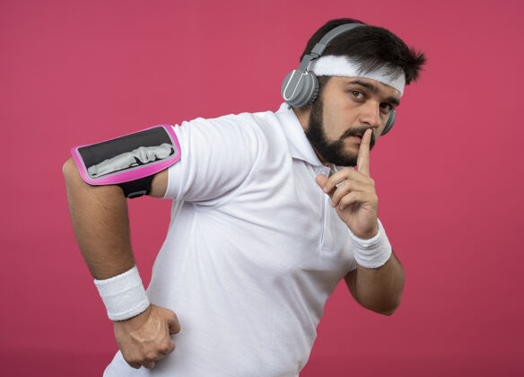 耳机年轻的运动型男子戴着头带和腕带 戴着耳机 手臂上戴着手机带 在粉红色的墙上显示出沉默的手势粉色年轻乐队