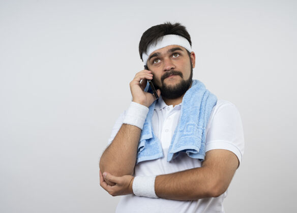 电话高兴的年轻运动男子抬头看戴着头带和手带与毛巾肩上讲电话隔离在白色的墙壁与复制空间男士腕带戴
