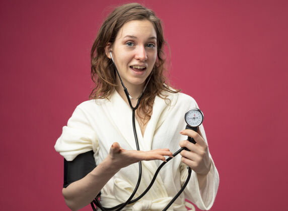 血压计高兴的年轻病女孩直视前方穿着白色长袍测量自己的压力血压计隔离在粉红色测量年轻压力