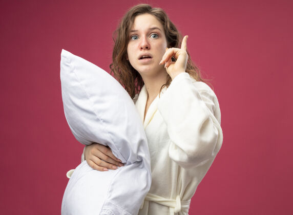 疾病印象深刻的年轻病女孩直直地向前看穿着白色长袍抱住枕头点在粉红色上孤立起来拥抱年轻枕头