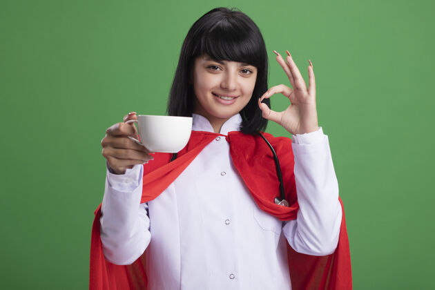 听诊器微笑着的年轻超级英雄女孩戴着听诊器 穿着医用长袍和斗篷 手里拿着一杯茶 在绿色的墙上显示出良好的姿态年轻斗篷女孩