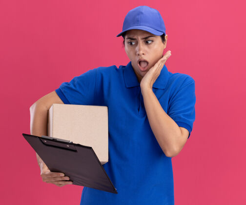 剪贴板年轻的送货员穿着制服 手里拿着帽子盒 惊讶地看着剪贴板 手放在粉红色墙上的脸颊上交货年轻举行