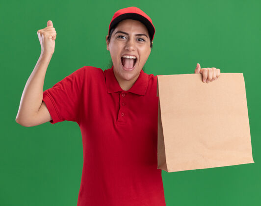 展示兴奋的年轻送货女孩穿着制服 戴着帽子拿着纸食品包 在绿色的墙上显示“是”的手势送货女孩抱着