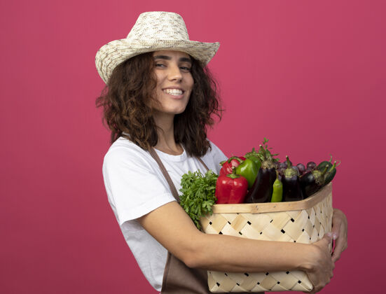人微笑的年轻女园丁穿着制服 戴着园艺帽 手里拿着一个粉红色的菜篮子年轻女人粉红