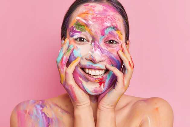 头像特写镜头正面的亚洲女人涂上了五颜六色的水粉颜料 手放在脸颊上笑得很开心深色女性脸颊