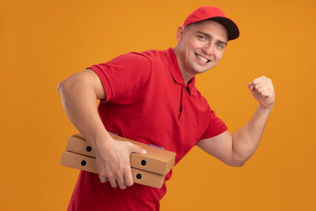 手势微笑的年轻送货员穿着制服 戴着帽子 拿着披萨盒 在橙色的墙上显示出强烈的手势强壮盒子年轻