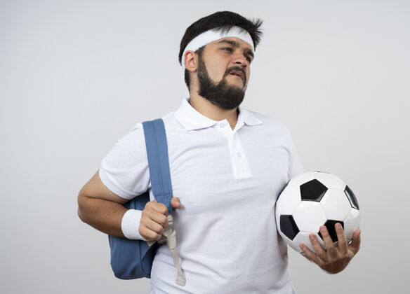 背包年轻的运动型男子戴着头带和腕带 背着背包 把球隔离在白色的墙上头带穿男子
