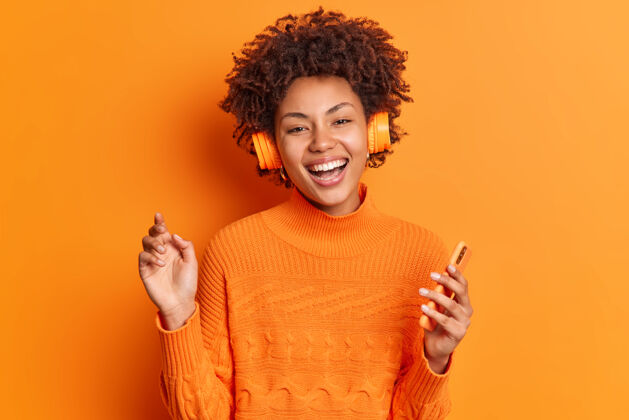 卷发人们娱乐和爱好的概念快乐的年轻非洲裔美国妇女卷发持有现代智能手机通过立体声耳机听音乐摆出生动的橙色背景套头衫年轻一个
