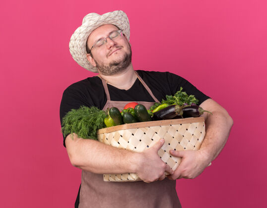 花园高兴地闭上眼睛的年轻男园丁戴着园艺帽 手里拿着菜篮子 孤零零地站在粉红色的墙上男性园艺封闭