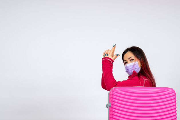 年轻女性正面图：戴着面具的粉色包的年轻女性在白墙上摆姿势度假大流行病毒女性covid-彩色旅行流行病微笑病毒