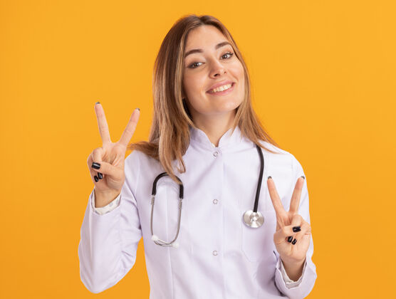 听诊器面带微笑的年轻女医生穿着医用长袍 听诊器在黄色的墙上显示出和平的姿态医生和平微笑