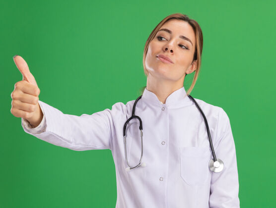 年轻高兴地看着身边年轻的女医生穿着医用长袍 听诊器显示拇指向上隔离在绿色的墙上医生听诊器壁板