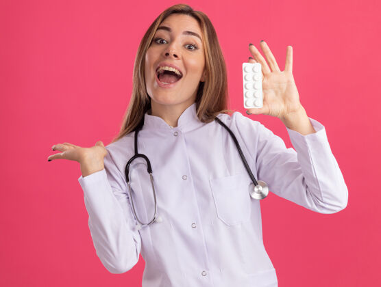 点兴奋的年轻女医生穿着医用长袍 手持听诊器 指着身后隔离的粉红色墙上的药丸听诊器药丸穿