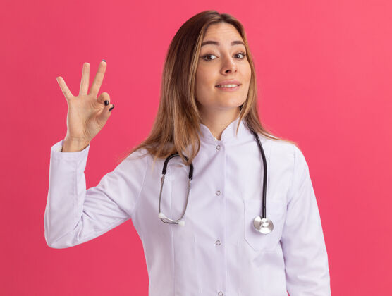 听诊器微笑的年轻女医生穿着医用长袍 听诊器显示出良好的姿态隔离在粉红色的墙上年轻表演微笑