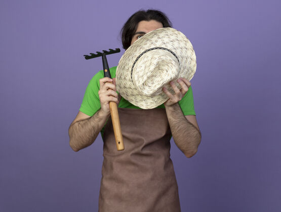 惊喜惊讶的年轻男园丁穿着制服拿着耙子蒙着脸 戴着紫色隔离帽耙脸花园