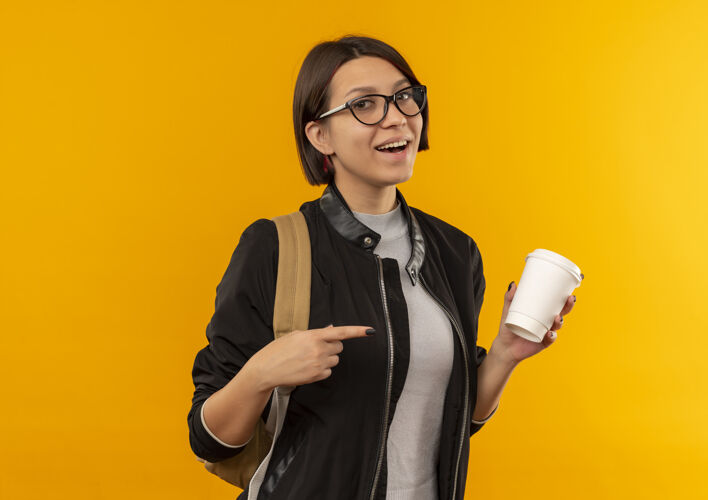 拿着快乐的年轻女学生戴着眼镜 背着书包 拿着塑料咖啡杯 指着孤立的橙色年轻眼镜杯子