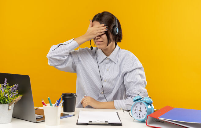 手年轻的呼叫中心女孩戴着耳机坐在办公桌旁 手里拿着工作工具 闭着眼睛 手被隔离在橙色背景上佩戴眼睛女孩