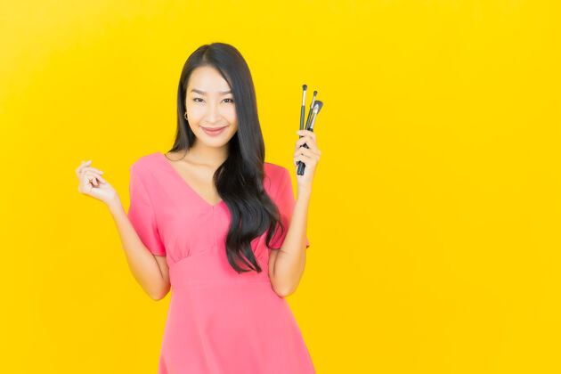 特写画像美丽的亚洲年轻女子微笑着用化妆刷在黄色的墙上清洁皮肤头发