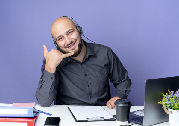 男人快乐的年轻秃头呼叫中心男子戴着耳机坐在办公桌旁 用工作工具做着呼叫手势 孤立在紫色上办公桌工具耳机