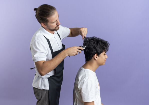 穿年轻帅气的理发师穿着制服站在侧视图中为年轻客户理发隔离在紫色背景上 留有复印空间制服年轻站立
