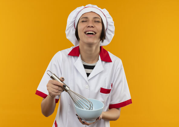 烹饪快乐的年轻女厨师穿着厨师制服拿着搅拌器和碗闭着眼睛孤立在橙色厨师持有眼睛