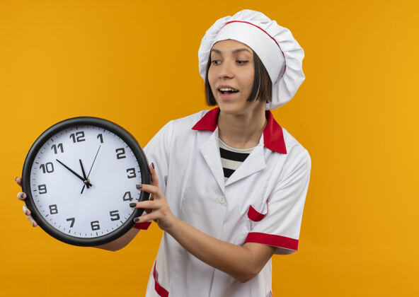 制服快乐的年轻女厨师穿着厨师制服 拿着桔子看着孤立的时钟快乐厨师钟