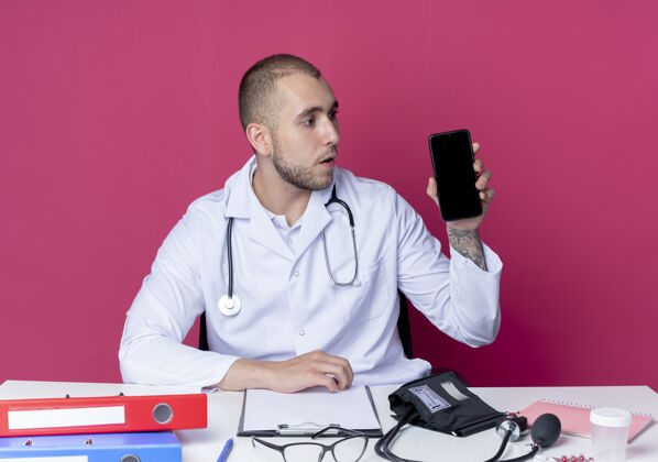 粉色印象深刻的年轻男医生穿着医用长袍和听诊器坐在办公桌前拿着工作工具 看着手机上孤立的粉红色长袍医生印象