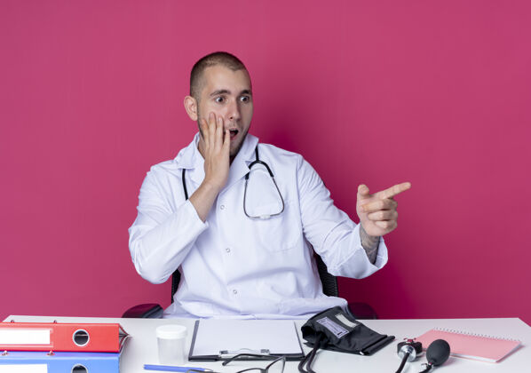 工作令人印象深刻的年轻男医生穿着医用长袍和听诊器坐在办公桌旁 手放在脸颊上 指着粉红色的一侧长袍工具壁板