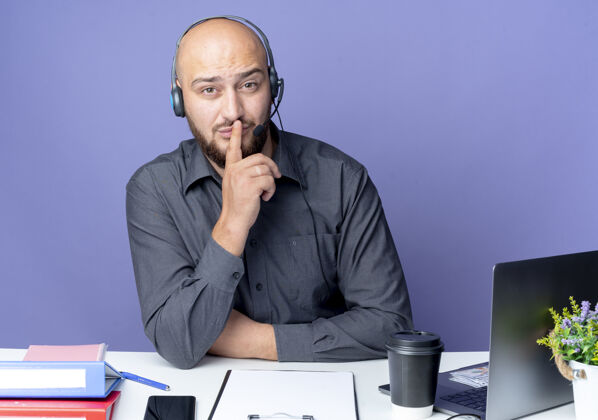 手势年轻秃顶的呼叫中心男子戴着耳机坐在办公桌旁 手里拿着工作工具 在紫色的背景下默默地打着手势中心秃头耳机