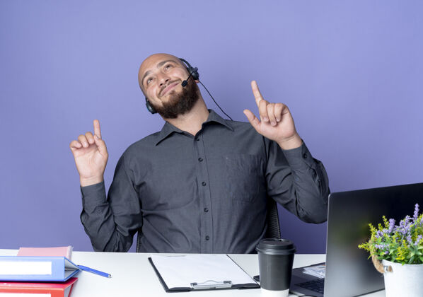 戴高兴的年轻秃顶呼叫中心男子戴着耳机坐在办公桌前 工作工具看着和指着紫色背景上孤立坐着指向中心