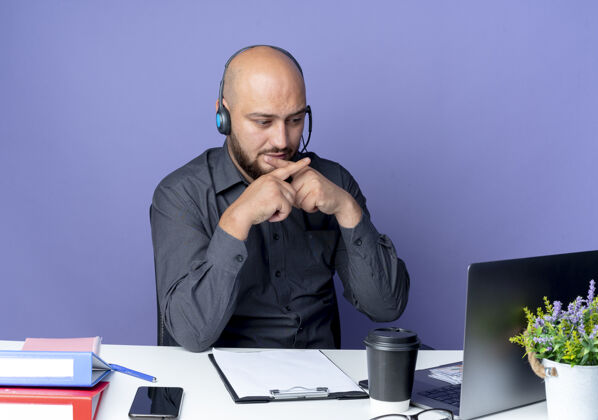 磨损戴着耳机的秃头呼叫中心年轻人坐在办公桌旁 手里拿着工作工具 看着笔记本电脑 在紫色的背景上做着不孤立的手势手势坐着中心