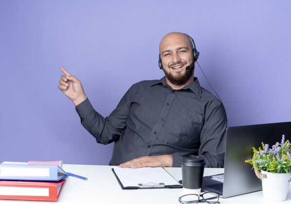 紫色快乐的年轻秃头呼叫中心男子戴着耳机坐在办公桌旁 工作工具指着紫色隔离的一侧壁板男人工具
