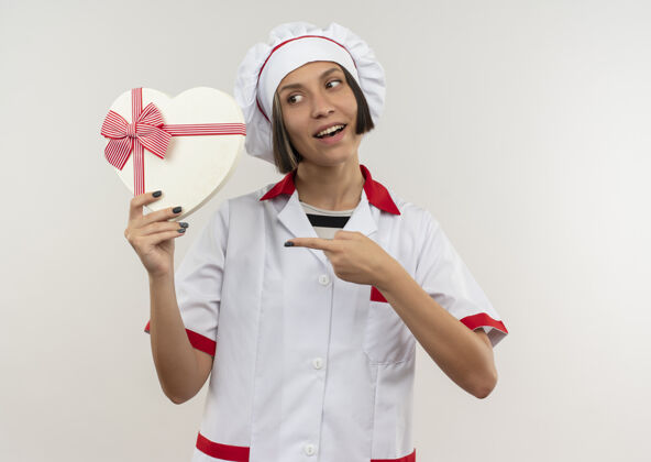 快乐身着厨师制服的快乐年轻女厨师手拿着一个白色的心形礼盒 看着并指着它厨师女拿着