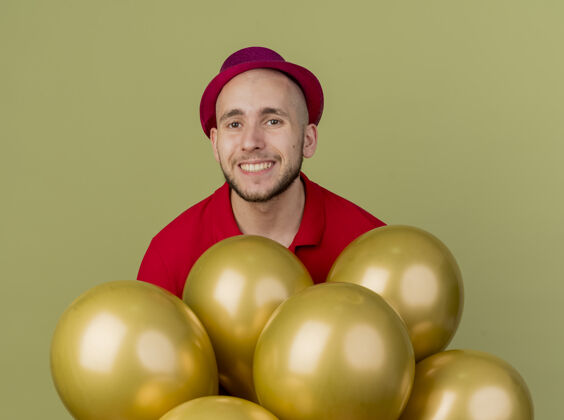 聚会微笑着的年轻英俊的斯拉夫党人戴着党的帽子站在气球后面看着前面孤立的橄榄绿墙上脸斯拉夫男人