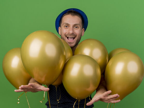 人戴着蓝色派对帽的快乐帅哥站在有复制空间的绿色墙上 手上伸出氦气球帅气姿势市民