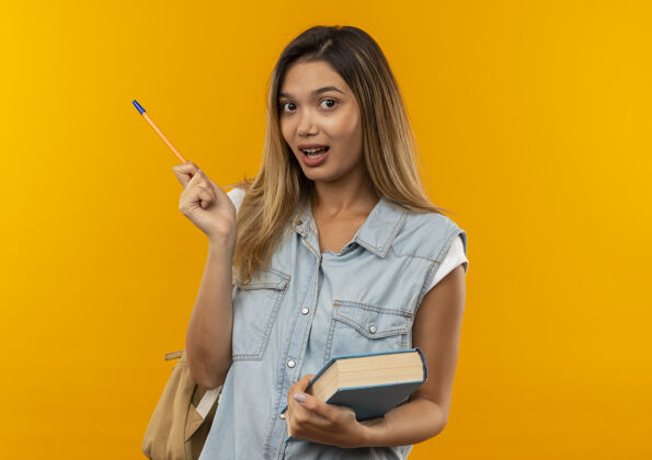 笔印象深刻的年轻漂亮的学生女孩穿着背袋举行的书和笔孤立的橙色年轻印象抱