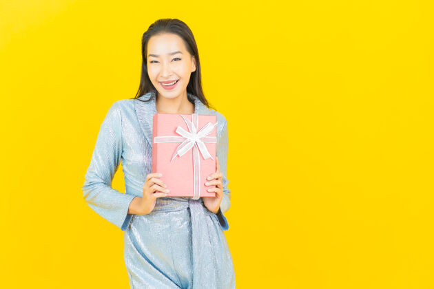 时尚肖像美丽的亚洲年轻女子微笑与红色礼品盒在黄色的墙壁节日微笑蓝色