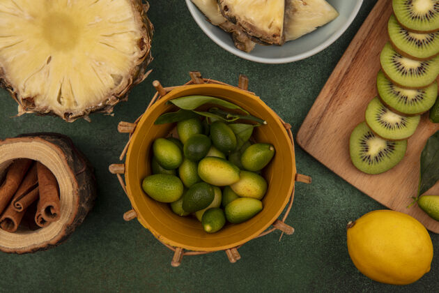 顶部桶上的芳香金盏花俯视图 木制厨房板上放着猕猴桃片 碗上放着菠萝 肉桂棒和柠檬隔离在绿色背景上菠萝视图木头