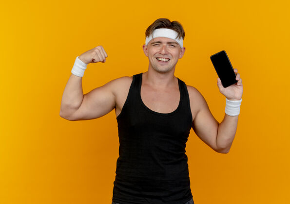男士快乐的年轻帅气的运动型男人戴着头带和腕带拿着手机 在橙色上做着强烈的孤立的手势橙色腕带佩戴