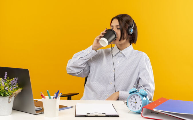 杯子年轻的呼叫中心女孩戴着耳机坐在办公桌旁 手里拿着工作工具 用塑料咖啡杯喝着咖啡 闭着眼睛隔离在橙色背景上电话女孩耳机