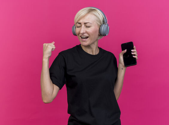 站着快乐的中年金发斯拉夫女人戴着耳机拿着手机做“是”的手势隔离在粉红色的墙上感觉女人金发