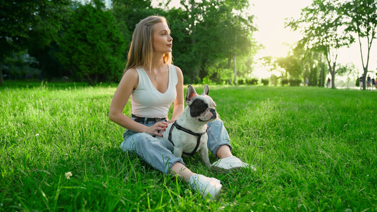 城市公园年轻快乐的女人和可爱的白色和棕色法国斗牛犬坐在新鲜的草地上的侧视图美丽微笑的女孩享受着夏日的夕阳 在城市公园爱犬人类和动物的友谊周末血统家庭
