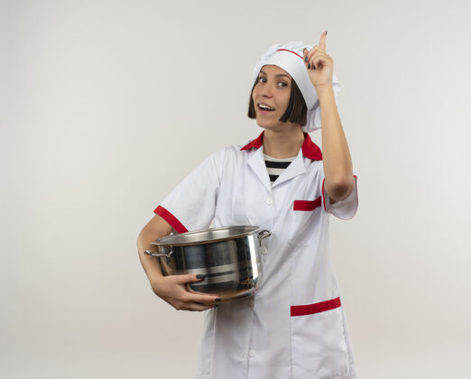 提高快乐的年轻女厨师穿着厨师制服拿着锅举起手指孤立在白色锅烹饪手指