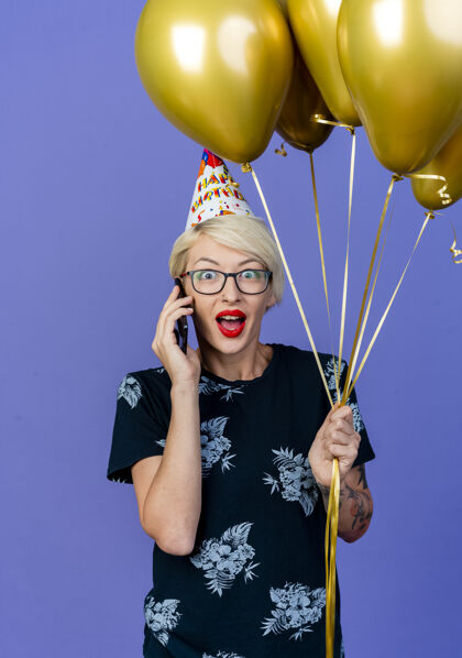 穿着令人印象深刻的年轻金发派对女子戴着眼镜和生日帽拿着气球讲电话看着前面孤立的紫色墙壁气球表情姿势
