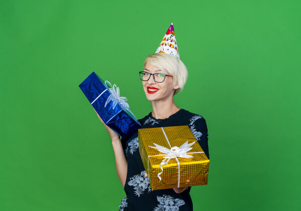 伸展微笑着的金发派对女郎戴着眼镜 戴着生日帽 拿着礼品盒 朝前伸展 看着隔离在绿色墙壁上的前面 还有复制空间人眼镜穿着