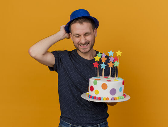 人高兴的帅哥戴着蓝色的派对帽 拿着生日蛋糕隔离在橙色的墙上 留着复制空间聚会请帅哥